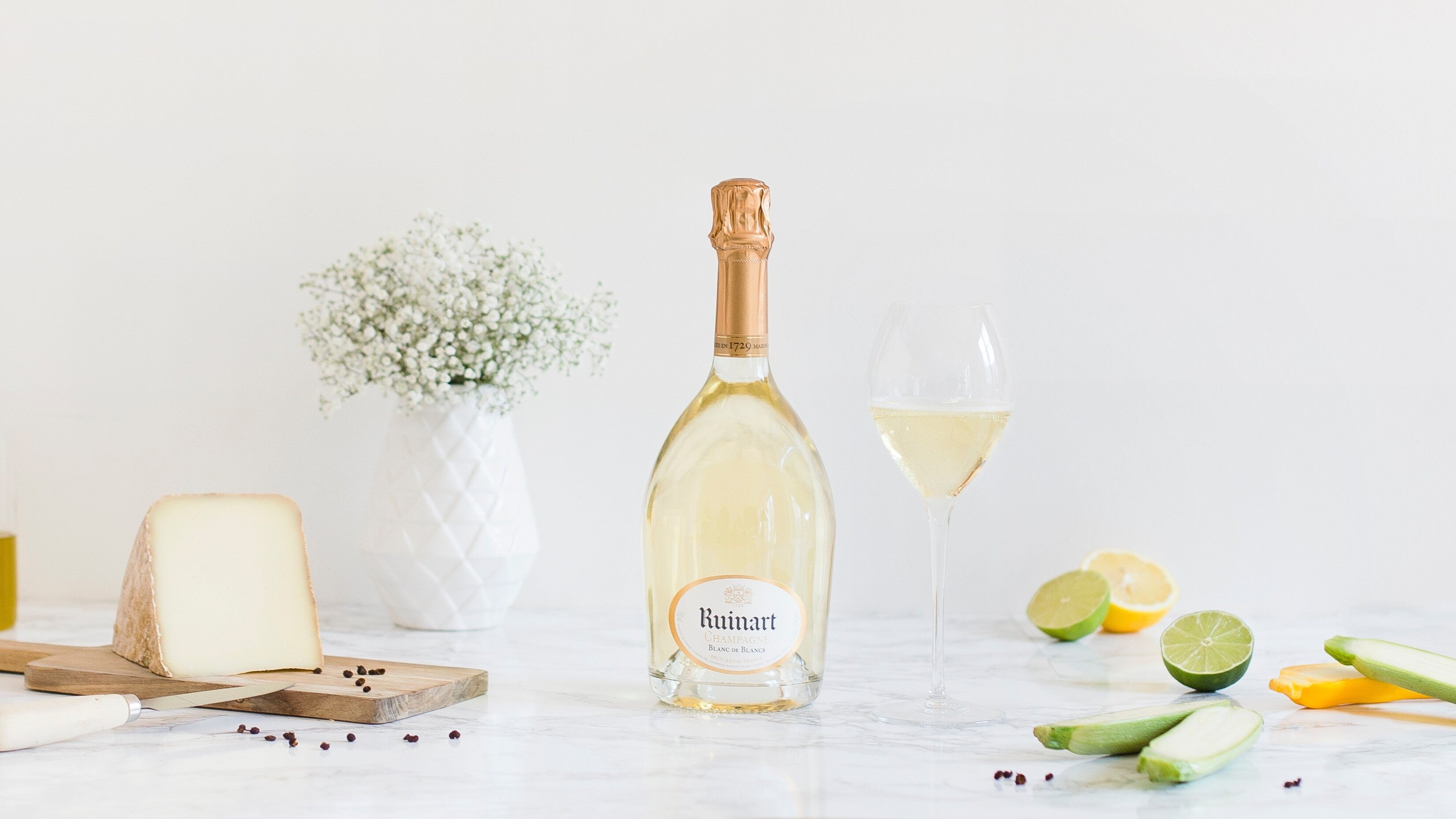 ottle of Ruinart Blanc de Blancs Champagne beside a filled wine glass, cheese, citrus, and vegetables on a bright tabletop.