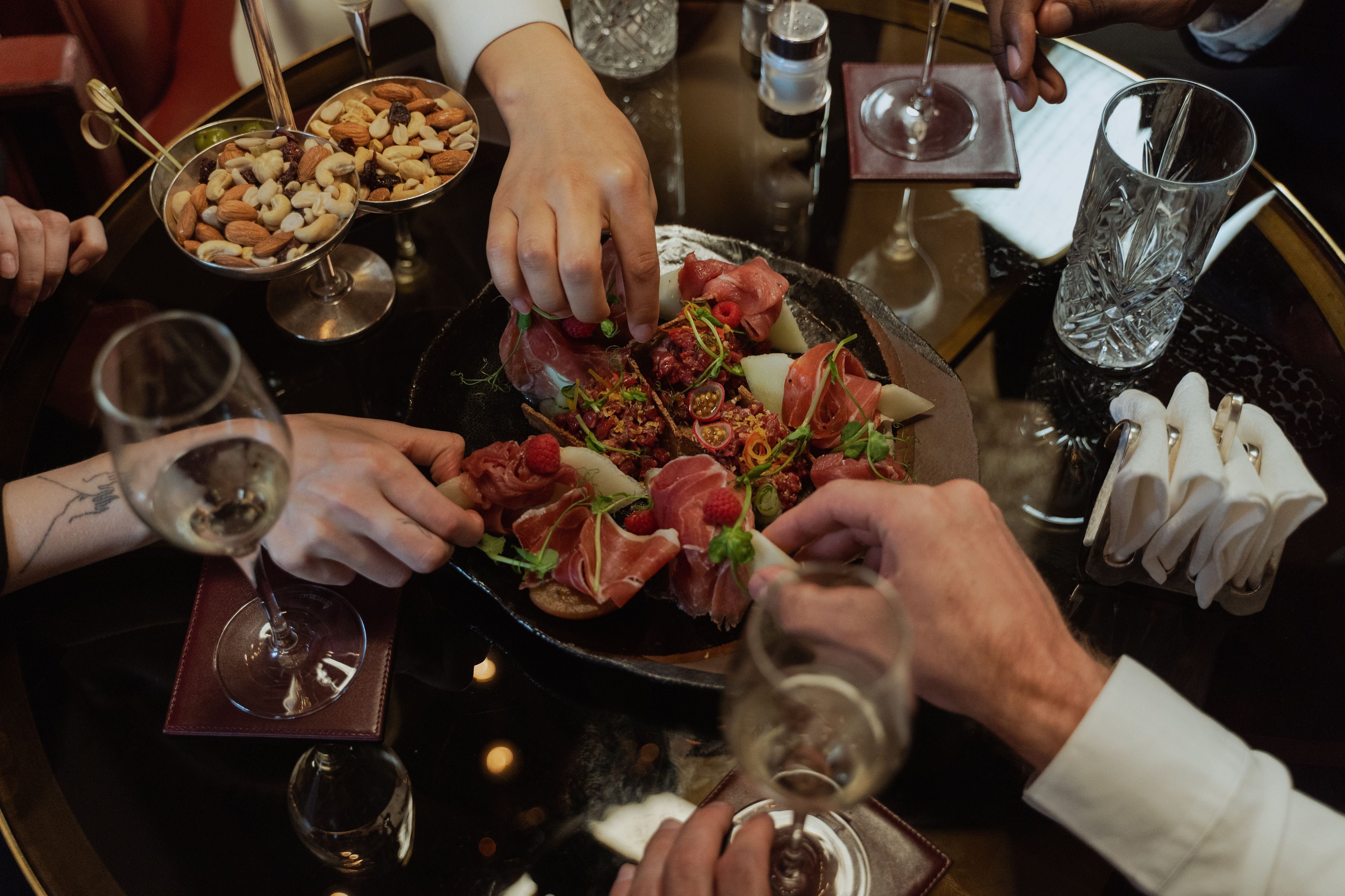 Guests share an elegant charcuterie platter with prosciutto, fruit, and cheese alongside wine glasses at a restaurant table.